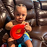 Josiah joined the competition — help win amazing prizes! baby, child, toy, red_ring, blue_shorts, braided_hair, leather_couch, indoor, playing, colorful_toys, person, sitting, furniture, cute, skin, hand, foot, young_child, expression, home