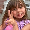 Nevaeh is registered to the contest to win money with this photo: child, girl, smile, peace_sign, hand, face, brown_hair, bangs, purple_shirt, outdoor, portrait, happy, cute, young, person, casual_clothing, expression, fingers, closeup, background_blur