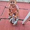 Lylah is registered to the contest to win money with this photo: toddler, child, baby, smiling, pink_outfit, hair_bows, sneakers, playground, outdoor, play_area, rubber_floor, toy, wheeled_toy, crouching, standing, fence, chain_link_fence, play_equipment, portrait, happy