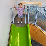 Lylah joined the competition — help win amazing prizes! toddler, child, indoor_playground, slide, smiling, standing, raised_hands, purple_shirt, shorts, sneakers, curly_hair, play_area, safety_gate, colorful, green_slide, wooden_wall, patterned_panels, portrait, recreation, happy