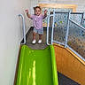 toddler, child, indoor_playground, slide, smiling, standing, raised_hands, purple_shirt, shorts, sneakers, curly_hair, play_area, safety_gate, colorful, green_slide, wooden_wall, patterned_panels, portrait, recreation, happy