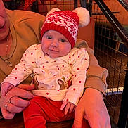 Lottie joined the competition — help win amazing prizes! baby, child, person, red_hat, knit_hat, pom_poms, holiday_clothing, festive, red_pants, white_shirt, stars_pattern, reindeer_design, adult_hands, jewelry, ring, wooden_table, christmas_decor, poinsettia, smile, indoor
