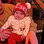 baby, child, person, red_hat, knit_hat, pom_poms, holiday_clothing, festive, red_pants, white_shirt, stars_pattern, reindeer_design, adult_hands, jewelry, ring, wooden_table, christmas_decor, poinsettia, smile, indoor