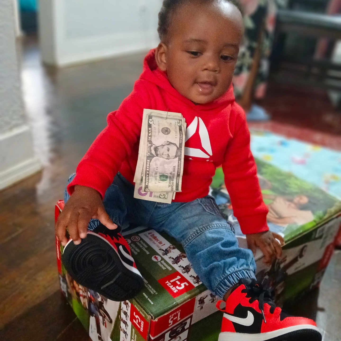 Trace'Ion is registered to the contest to win money with this photo: box, casual, child, clothing, curious, cute, dollar_bills, footwear, home, indoor, jeans, money, person, portrait, red_hoodie, sitting, sneakers, toddler, wooden_floor, young_child
