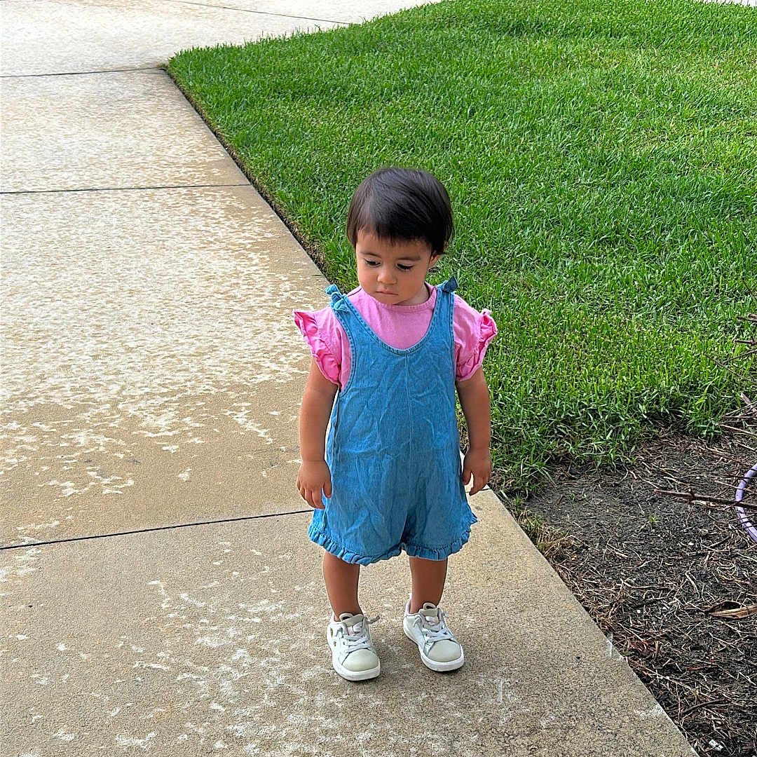 Natalia is registered to the contest to win money with this photo: toddler, child, sidewalk, grass, outdoor, greenery, building, stairs, footwear, clothing, shorts, person, curly_hair, daylight, casual, pink, blue, shoes, nature, concrete