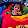 Aurora is registered to the contest to win money with this photo: baby, child, plush_toy, red, yellow, green, blue, car_seat, indoor, person, face, tongue_out, hands, furniture, fan, room, clutter, toy, soft, cute