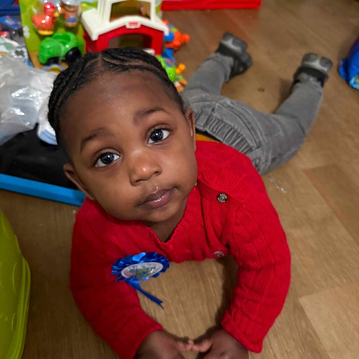 Kartier is registered to the contest to win money with this photo: child, toddler, red_sweater, indoor, wooden_floor, toys, playroom, looking_up, curious, baby, hair_braids, blue_ribbon, casual_clothing, floor, hands, eyes, face, person, home, play