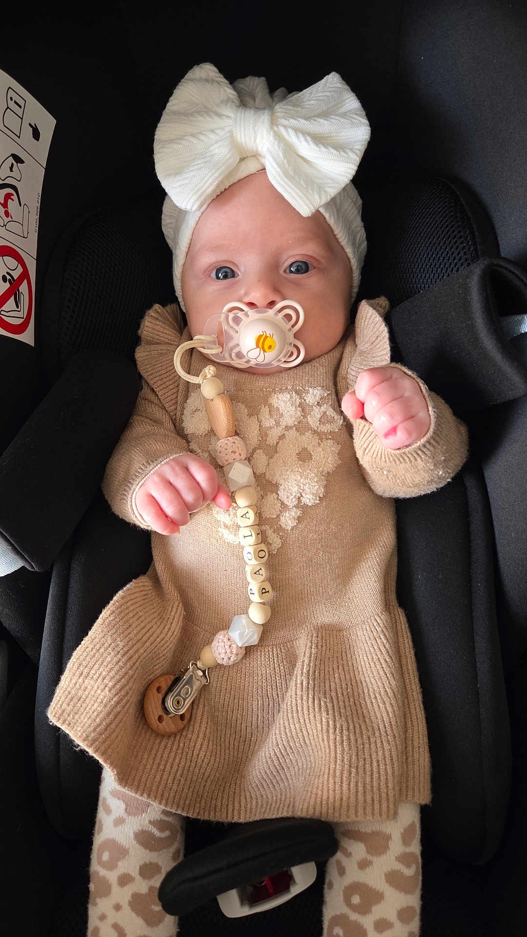 Paola participe au concours pour gagner de l'argent avec cette photo : baby, bow, pacifier, car_seat, knitted_dress, beige, patterned_tights, pacifier_clip, name_beads, infant, child, cute, portrait, indoors, seat_belt, clothing, hands, headwear, face, person