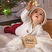 Elara is registered to the contest to win money with this photo: baby, blanket, celebration, child, christmas_tree, clothing, curious, cute, decorations, festive, floor, hat, holiday, home, indoor, infant, lights, ornament, reaching, toddler