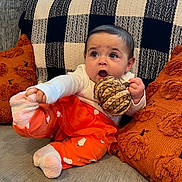 Elara is registered to the contest to win money with this photo: baby, child, infant, couch, pillow, plush, toy, orange_clothing, socks, cute, expression, indoors, home, comfort, seated, playful, texture, blanket, face, person