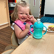 Ayla is registered to the contest to win money with this photo: toddler, child, smiling, pink_bib, sippy_cup, wooden_table, plate, food, indoor, classroom, chair, floor, door, cabinet, happy, person, young_child, casual, daylight, cute