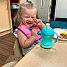 toddler, child, smiling, pink_bib, sippy_cup, wooden_table, plate, food, indoor, classroom, chair, floor, door, cabinet, happy, person, young_child, casual, daylight, cute