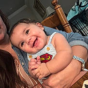 Colson joined the competition — help win amazing prizes! baby, child, smile, drool, person, superman_logo, clothing, indoor, adult, hand, table, chair, wood, watch, jewelry, happy, face, holding, casual, long_hair