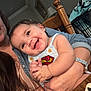 baby, child, smile, drool, person, superman_logo, clothing, indoor, adult, hand, table, chair, wood, watch, jewelry, happy, face, holding, casual, long_hair