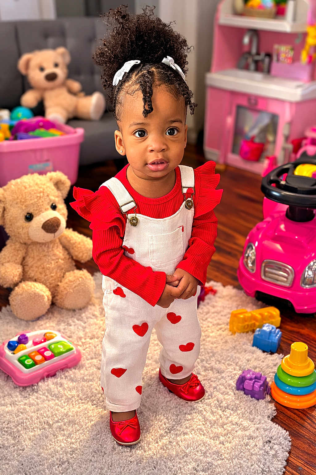 Lynn Hayley is registered to the contest to win money with this photo: child, toddler, girl, curly_hair, white_bows, red_shoes, heart_overalls, stuffed_teddy, pink_toy_car, stacking_rings, playroom, rug, wood_floor, toy_piano, portrait, big_eyes, cute, smile, standing, indoor