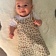 Hallie is registered to the contest to win money with this photo: baby, infant, smiling, face, leopard_print, dress, white_shirt, buttons, soft_rug, lying_down, cute, child, apparel, happy, skin, head, eyes, hand, footwear, indoors