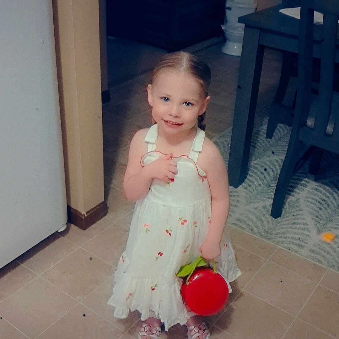 Cambria is registered to the contest to win money with this photo: child, girl, dress, white_dress, braided_hair, smiling, red_bucket, indoor, kitchen, floor_tiles, dining_table, chairs, carpet, toy, happy, cute, portrait, young_child, love_text, black_heart_emoji
