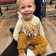 Maverick is registered to the contest to win money with this photo: child, smiling, sitting, floor, shirt, text, shoes, pants, indoor, people, legs, wood_floor, chair, casual_clothing, happy, young, person, face, hair, light