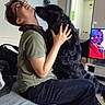 affection, black_dog, black_hair, casual_clothing, couch, dog, embrace, furniture, glasses, happy, home, human_dog_interaction, indoor, living_room, person, pet, sitting, smiling, technology, television