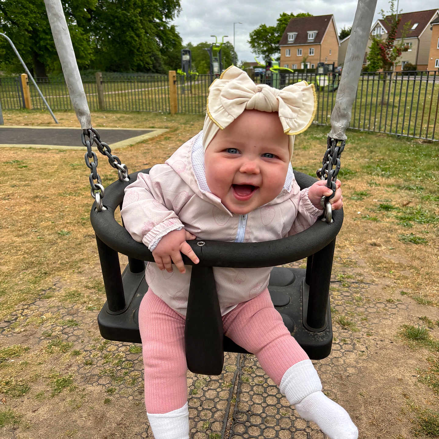 Lucia Rose is registered to the contest to win money with this photo: baby, bow, child, clothing, cloudy_sky, fence, grass, happy, headband, houses, jacket, metal_chains, outdoor, pink_pants, playground, seat, smile, swing, trees, white_socks