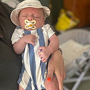 Austin is registered to the contest to win money with this photo: baby, sleeping, pacifier, hat, striped_clothing, hand, adult, nail_polish, orange_nail_polish, romper, infant, closeup, indoor, person, holding, cute, newborn, soft_lighting, peaceful, portrait