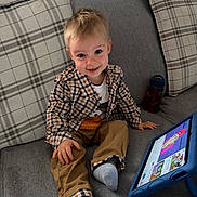 Elliot is registered to the contest to win money with this photo: child, clothing, couch, face, furniture, hair, happy, home, indoor, pants, person, plaid_pillow, shirt, sitting, smiling, socks, tablet, technology, toddler, water_bottle