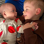 Edison is registered to the contest to win money with this photo: baby, toddler, siblings, children, portrait, close_up, smiling, lying_down, affection, interaction, face, eyes, hands, apple_print, clothing, brown_shirt, blue_background, couch, indoors, family