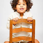 Yolann a rejoint le concours — aidez-le/la à gagner de superbes lots ! casual_clothing, child, curly_hair, cute, face, hands, happy, indoors, long_sleeve_shirt, necklace, person, portrait, shorts, sitting, smiling, studio, toddler, white_background, wooden_chair, young_child
