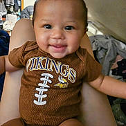 Zoey joined the competition — help win amazing prizes! baby, infant, child, smiling, portrait, indoors, bedroom, held, sports_jersey, football, team_logo, brown_clothing, sitting, closeup, face, eyes, hair, arms, home, happy