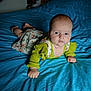 baby, infant, bed, blue_bedspread, floral_dress, green_sweater, hand, face, person, indoor, furniture, blanket, sleepwear, child, skin, head, expression, lying_down, pale_skin, curious