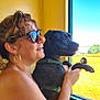 woman, dog, black_dog, sunglasses, window, sunlight, outdoor_view, green_field, happy, smiling, curly_hair, earrings, pet, human_animal_interaction, leash, holding, looking_out, daylight, portrait, casual