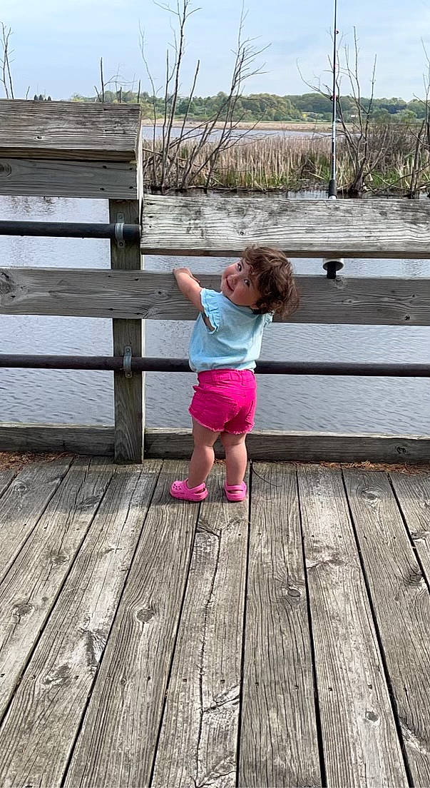 Camilla is registered to the contest to win money with this photo: bank, boardwalk, dock, fence, grass, happy, hardwood, human_leg, lake, leisure, people_in_nature, person, plank, plant, sky, standing, toddler, tree, water, wood