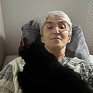 Solange Fournou a rejoint le concours — aidez-le/la à gagner de superbes lots ! animal, black_cat, casual, cat, closeup, comfort, couch, cozy, glasses, home, indoor, knitted, older_adult, pet, pillow, portrait, relaxing, resting, sweater, woman