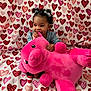 Nova is registered to the contest to win money with this photo: baby, child, curly_hair, bows, pink_teddy_bear, plush_toy, heart_pattern, blanket, cute, indoor, smiling, chewing, cozy, soft_toy, happy, infant, seated, colorful, playful, adorable