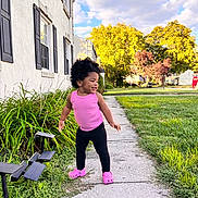 Nova is registered to the contest to win money with this photo: toddler, child, sidewalk, pink_clothing, curly_hair, greenery, house, windows, plants, grass, outdoor, sunny, trees, clouds, residential_area, footwear, crocs, smiling, happy, person