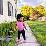 toddler, child, sidewalk, pink_clothing, curly_hair, greenery, house, windows, plants, grass, outdoor, sunny, trees, clouds, residential_area, footwear, crocs, smiling, happy, person