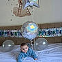 baby, balloon, banner, bed, birthday, blue_sweater, celebration, child, cute, decorations, festive, happy, indoor, infant, lying_down, party_hat, room, silver_balloon, smile, toddler
