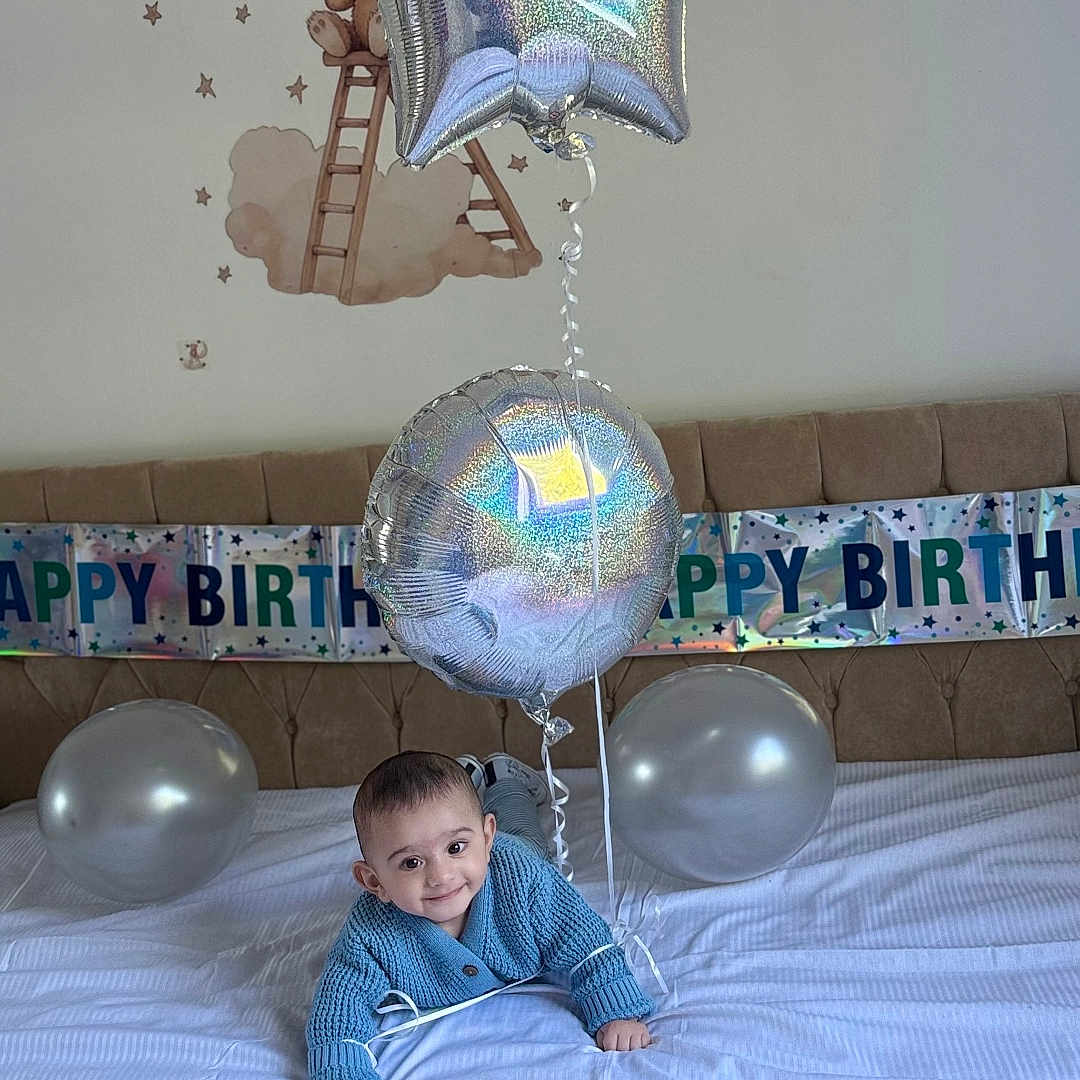 Zuhayr is registered to the contest to win money with this photo: baby, balloon, banner, bed, birthday, blue_sweater, celebration, child, cute, decorations, festive, happy, indoor, infant, lying_down, party_hat, room, silver_balloon, smile, toddler