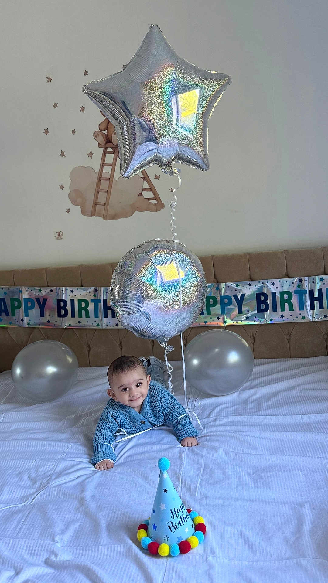 Zuhayr is registered to the contest to win money with this photo: baby, child, birthday, balloon, silver_balloon, party_hat, blue_sweater, bed, smile, celebration, happy, indoor, decorations, banner, festive, infant, cute, lying_down, room, toddler