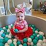 baby, child, ball_pit, balls, red_dress, headband, pink_bow, smile, indoor, flooring, furniture, wooden_chair, baby_seat, toy_mobile, face, feet, person, cute, happy, toddler