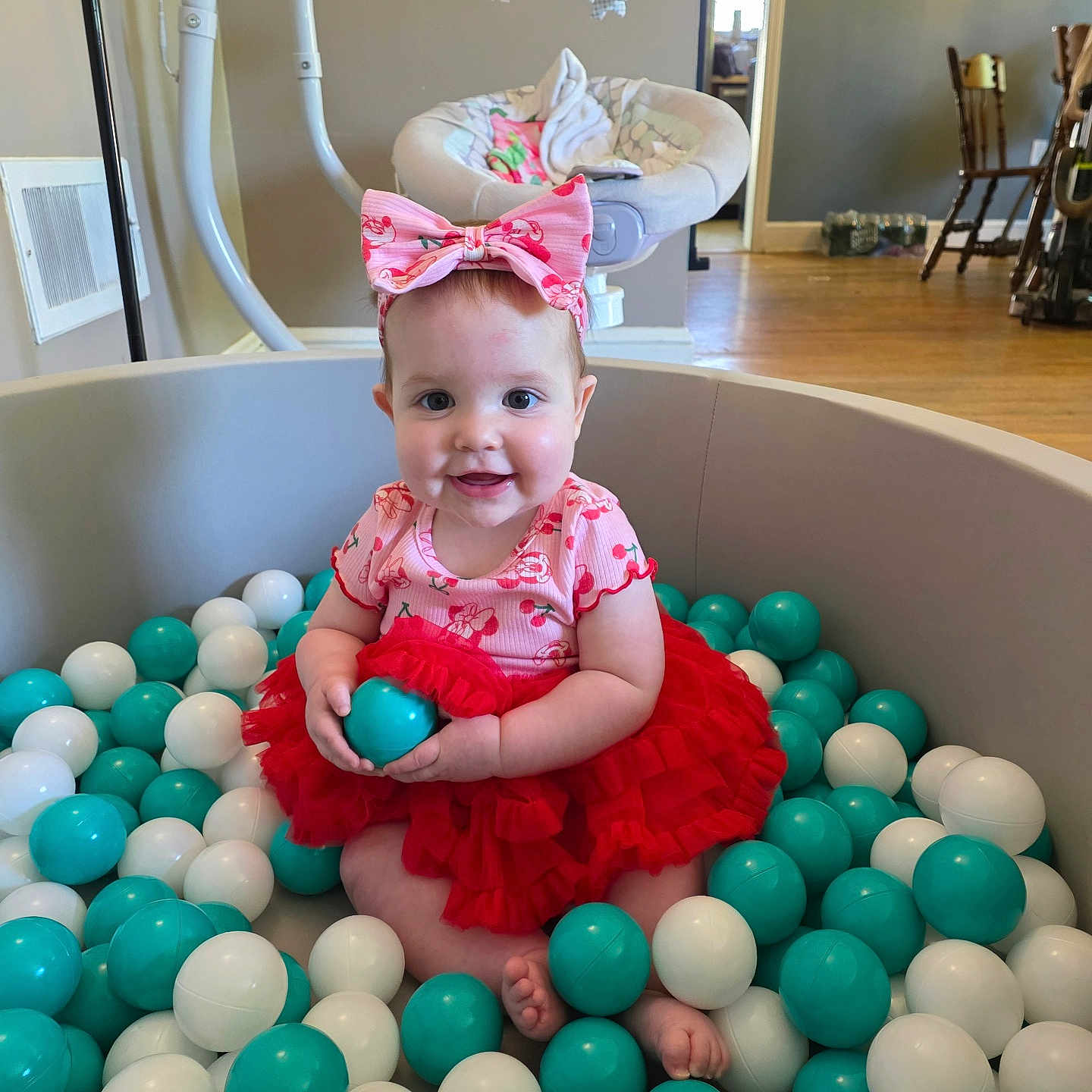 Taytum is registered to the contest to win money with this photo: baby, child, ball_pit, balls, red_dress, headband, pink_bow, smile, indoor, flooring, furniture, wooden_chair, baby_seat, toy_mobile, face, feet, person, cute, happy, toddler