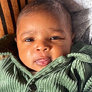 Zarah a rejoint le concours — aidez-le/la à gagner de superbes lots ! baby, infant, face, closeup, green_jacket, ribbed_fabric, eyes, mouth, skin, hand, patterned_background, warm_lighting, cozy, clothing, portrait, young_child, cute, indoor, soft_texture, expression