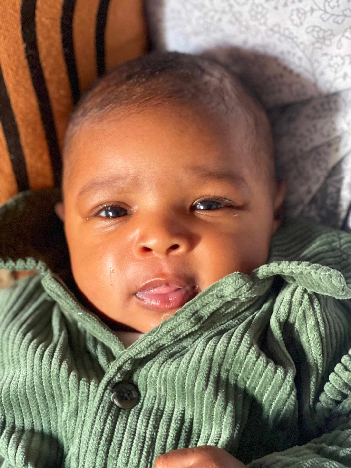 Zarah a rejoint le concours — aidez-le/la à gagner de superbes lots ! baby, infant, face, closeup, green_jacket, ribbed_fabric, eyes, mouth, skin, hand, patterned_background, warm_lighting, cozy, clothing, portrait, young_child, cute, indoor, soft_texture, expression