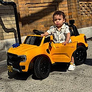 Hasiel is registered to the contest to win money with this photo: toddler, child, toy_car, yellow_car, outdoor, sidewalk, brick_wall, sunlight, shadow, shoes, shirt, curly_hair, small_vehicle, playtime, street, urban, daylight, door_open, expression, hand_on_car