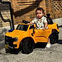 toddler, child, toy_car, yellow_car, outdoor, sidewalk, brick_wall, sunlight, shadow, shoes, shirt, curly_hair, small_vehicle, playtime, street, urban, daylight, door_open, expression, hand_on_car