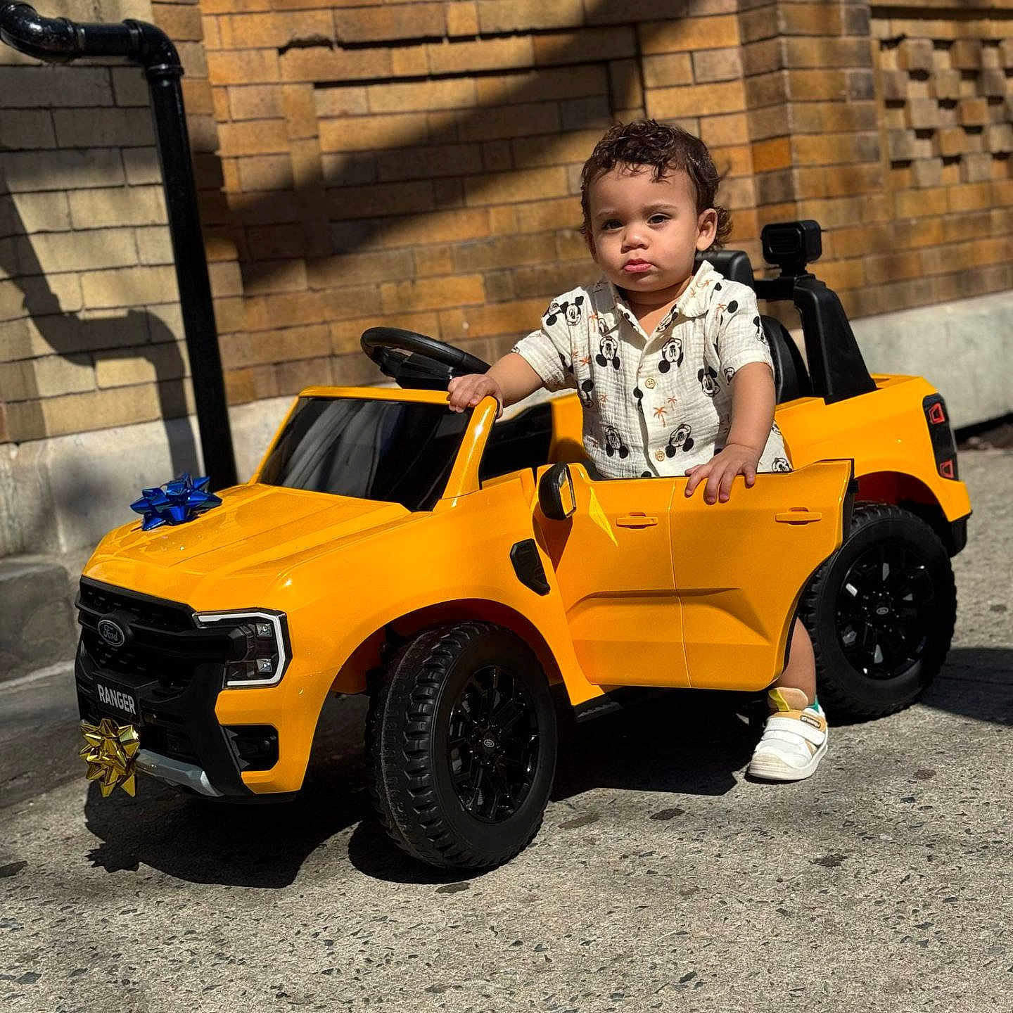 Hasiel is registered to the contest to win money with this photo: brick_wall, child, curly_hair, daylight, door_open, expression, hand_on_car, outdoor, playtime, shadow, shirt, shoes, sidewalk, small_vehicle, street, sunlight, toddler, toy_car, urban, yellow_car