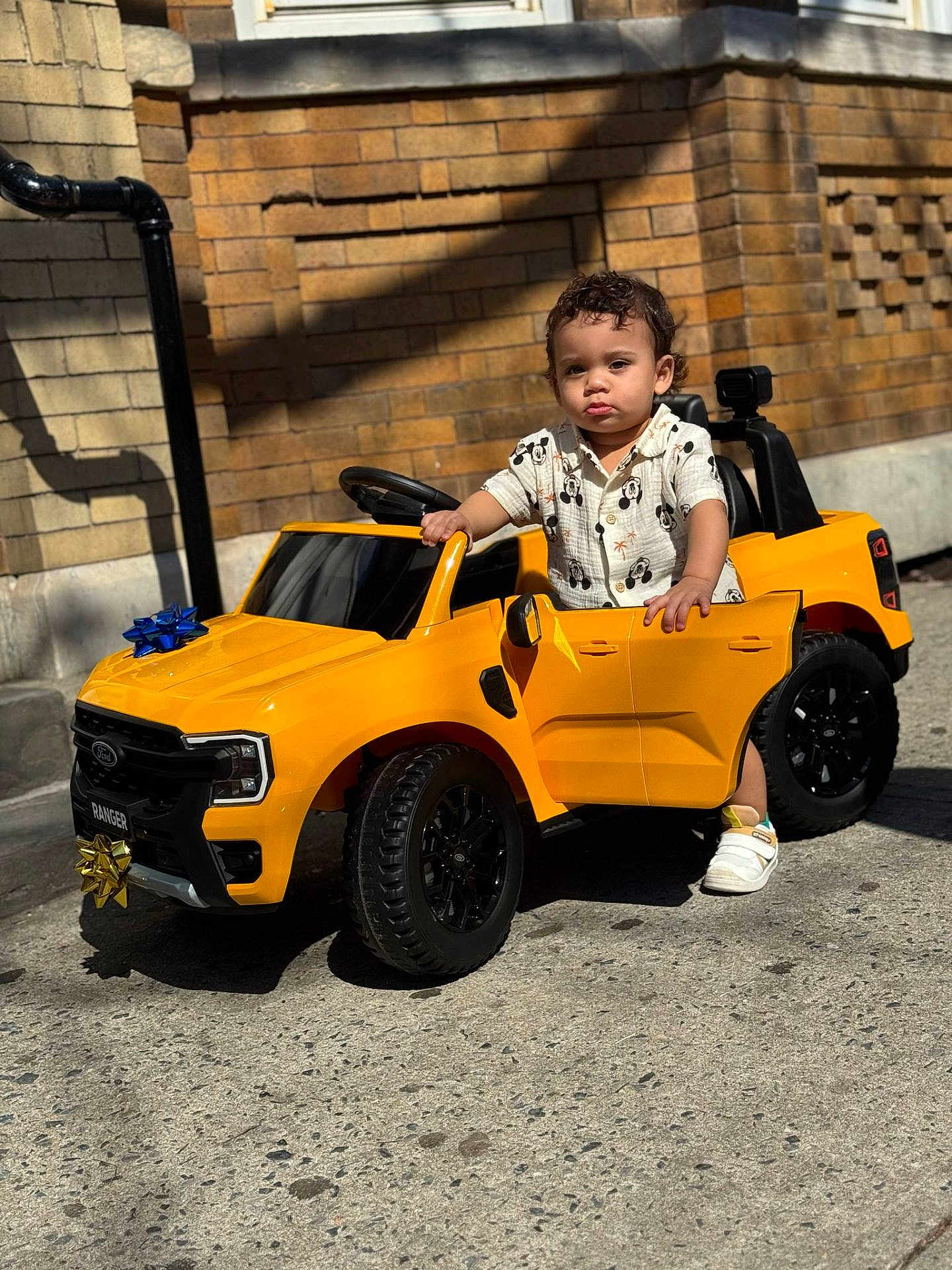 Hasiel is registered to the contest to win money with this photo: toddler, child, toy_car, yellow_car, outdoor, sidewalk, brick_wall, sunlight, shadow, shoes, shirt, curly_hair, small_vehicle, playtime, street, urban, daylight, door_open, expression, hand_on_car