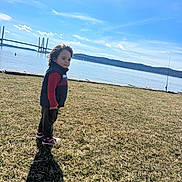 Hasiel is registered to the contest to win money with this photo: bridge, calm, child, clothing, daylight, grass, hill, landscape, nature, outdoor, person, red_shirt, scenic, shadow, sky, sneakers, standing, thoughtful, vest, water