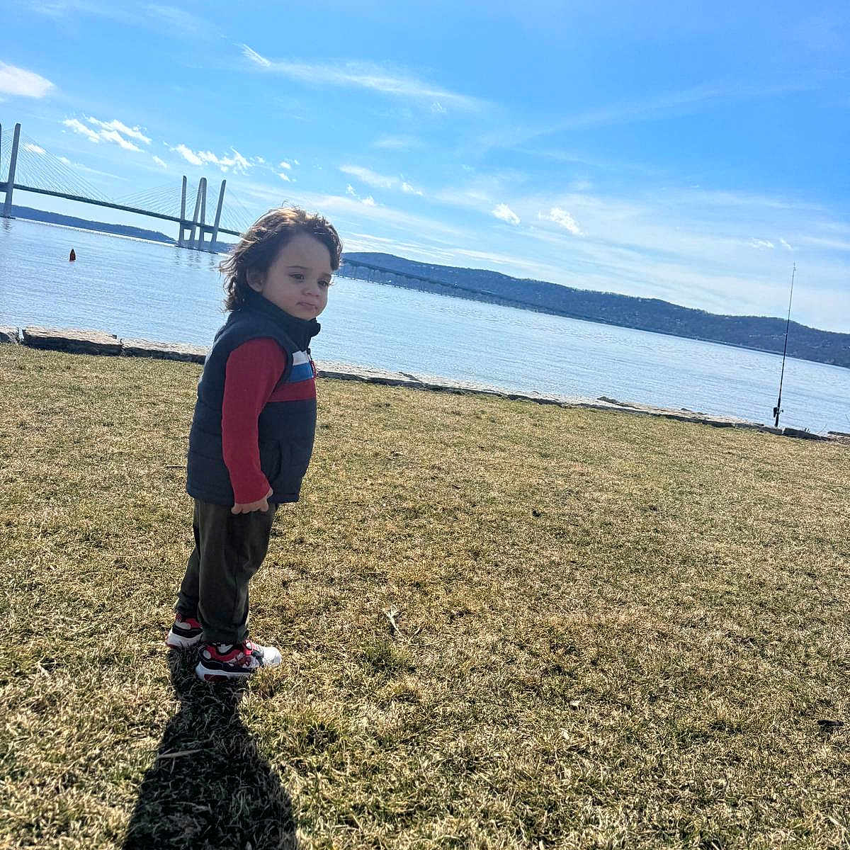 Hasiel is registered to the contest to win money with this photo: bridge, calm, child, clothing, daylight, grass, hill, landscape, nature, outdoor, person, red_shirt, scenic, shadow, sky, sneakers, standing, thoughtful, vest, water