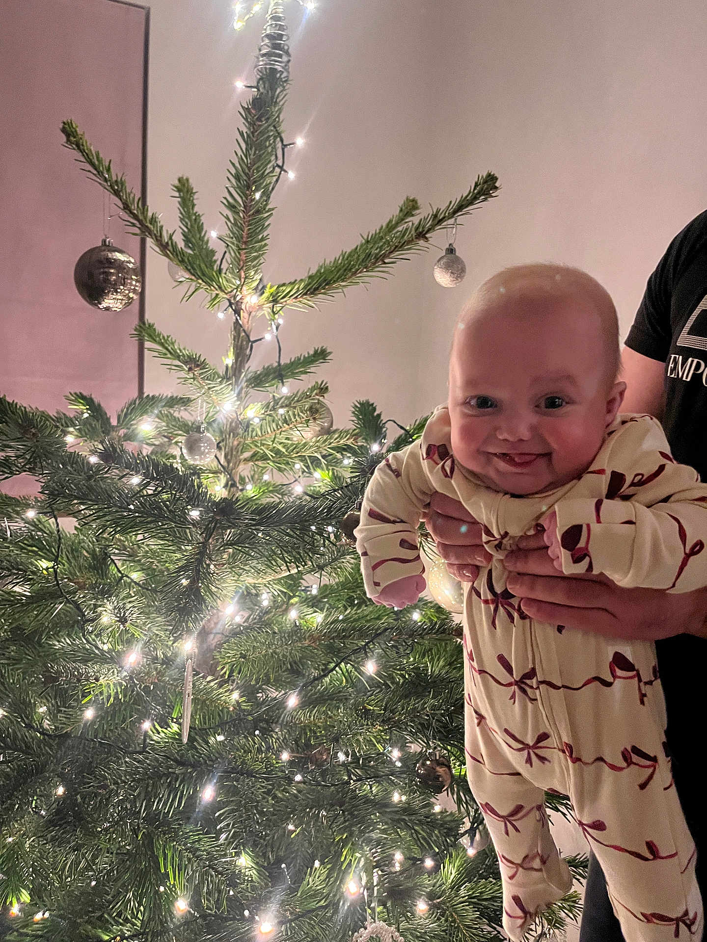 Hallie is registered to the contest to win money with this photo: baby, child, christmas_tree, lights, ornaments, holiday, festive, pajamas, smile, person, indoor, celebration, greenery, hands, joyful, decoration, tree, human, holding, cheerful
